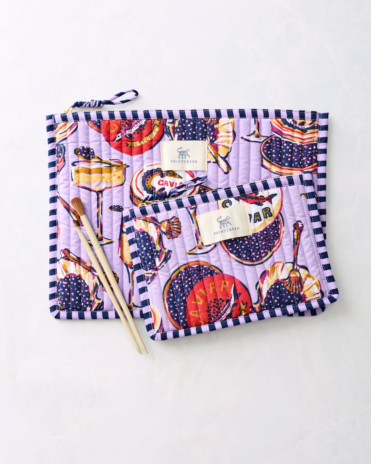 Two lavender Champagne & Caviar flat pouches with striped trim and zipper closures, shown with chopsticks.