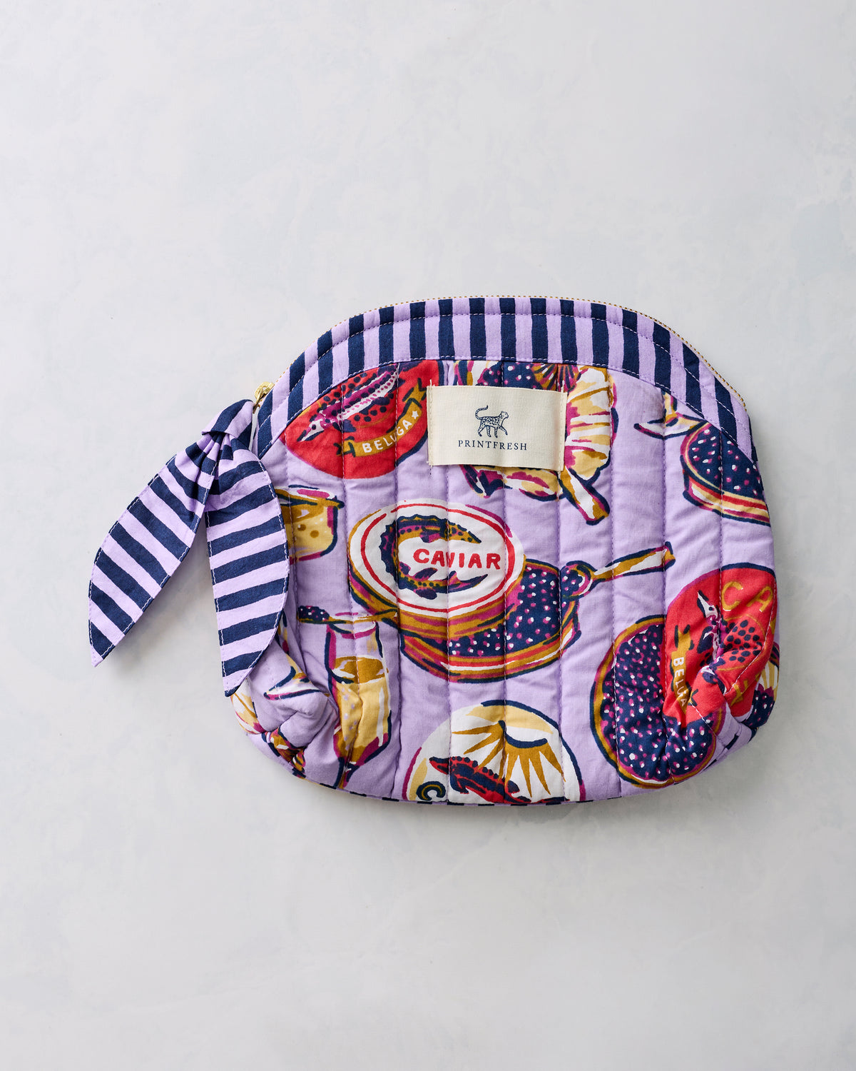Lavender quilted Champagne & Caviar makeup bag with rounded top, striped trim, zipper closure, and side tie.