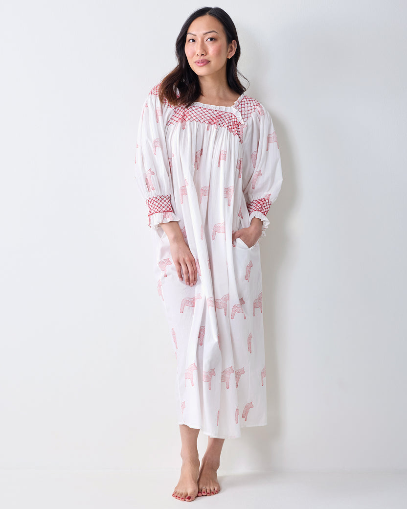 Front view of a person wearing a long white nightgown with pink horse prints, red smocked trim, and side pockets.