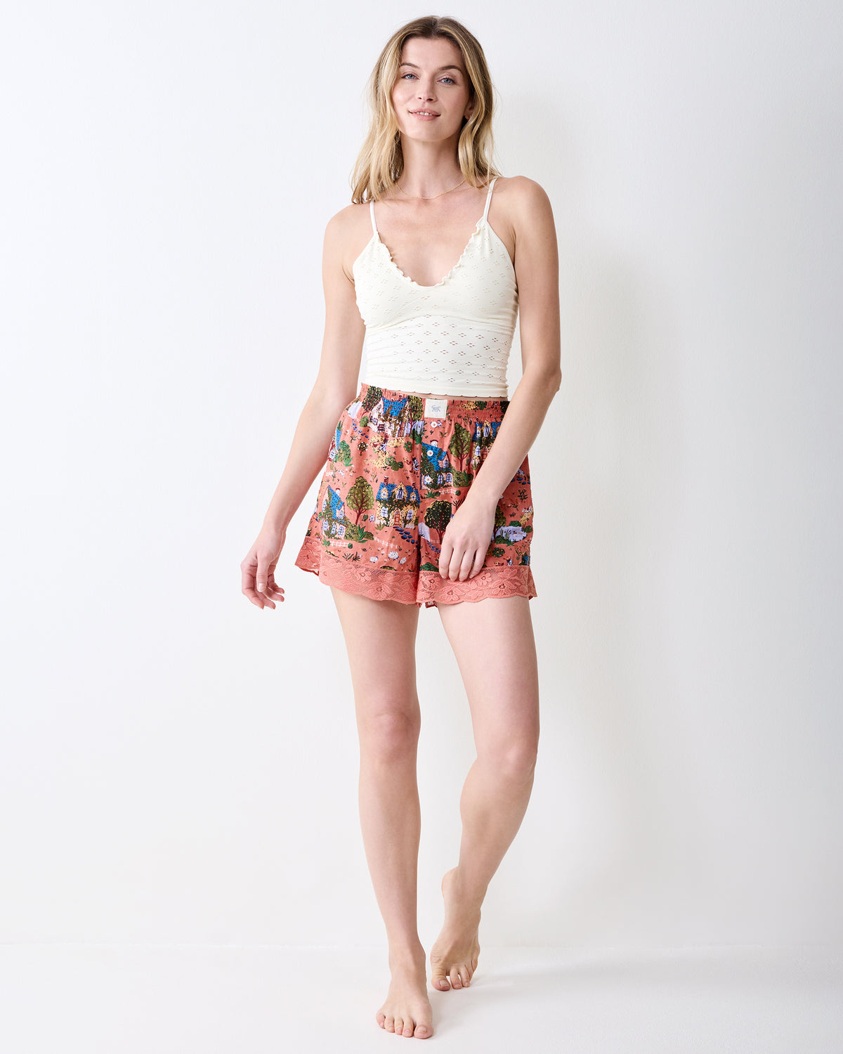 Front view of dusty rose pajama shorts with a cottage-and-tree print and lace-trim hems, worn with a white camisole.