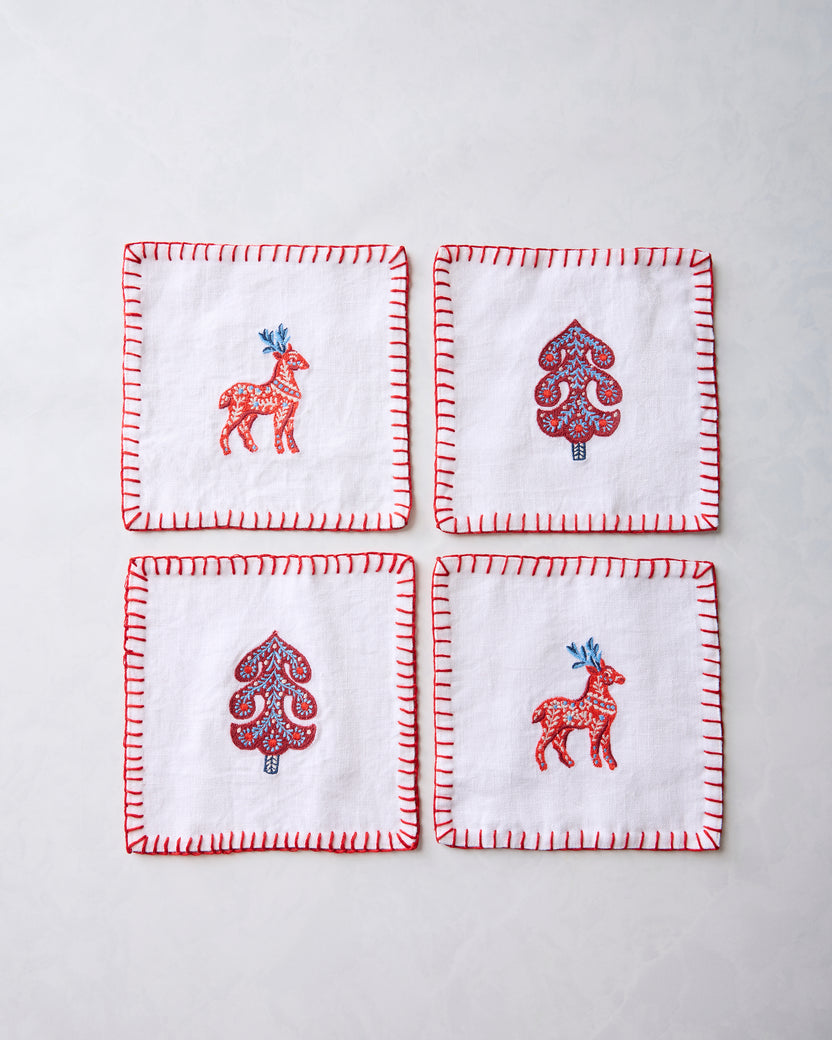 Four white cocktail napkins arranged in a square, each with red stitched edges and an embroidered deer or stylized tree motif.