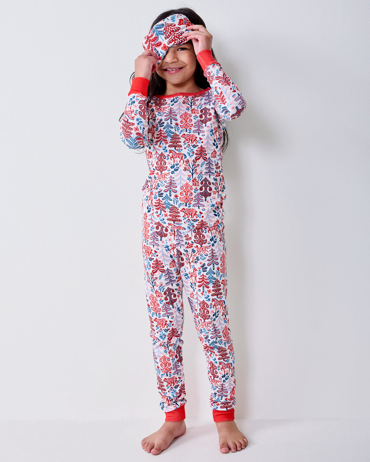 Child wearing a cream long-sleeve pajama top and pants with a red and blue Folklore Forest print, holding a matching sleep mask.