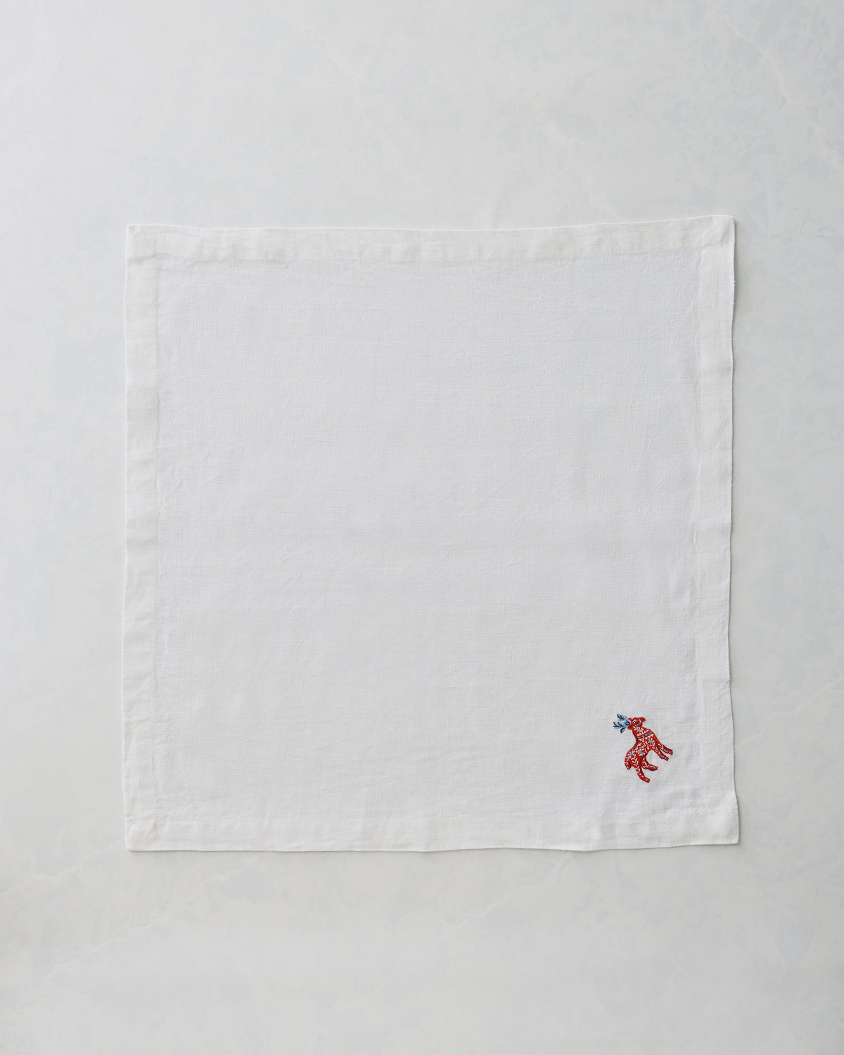 White oversized dinner napkin shown flat, with a small embroidered red deer and blue antlers near the lower-right corner.