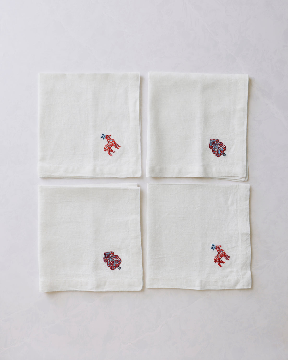 Four folded white oversized dinner napkins arranged in a square, with small embroidered deer and stylized tree motifs near the corners.