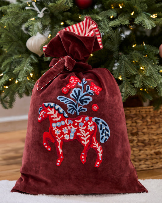 Upright burgundy gift sack with a red embroidered horse and floral motif, shown beside a decorated Christmas tree.