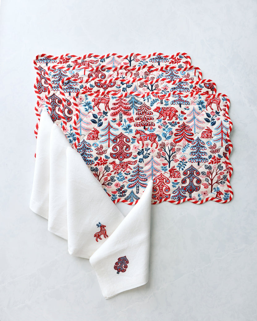 Stacked patterned placemats with white dinner napkins, featuring red, blue, and pink forest motifs.