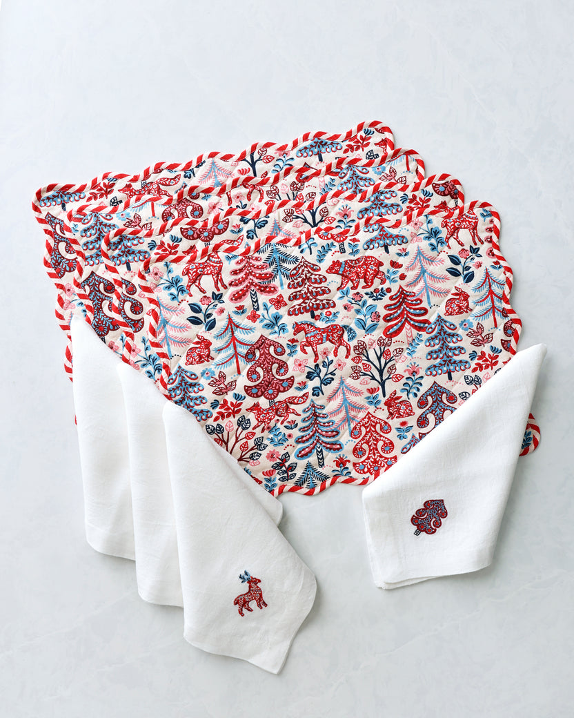 Four patterned placemats layered with white dinner napkins, showing red, blue, and pink forest motifs.