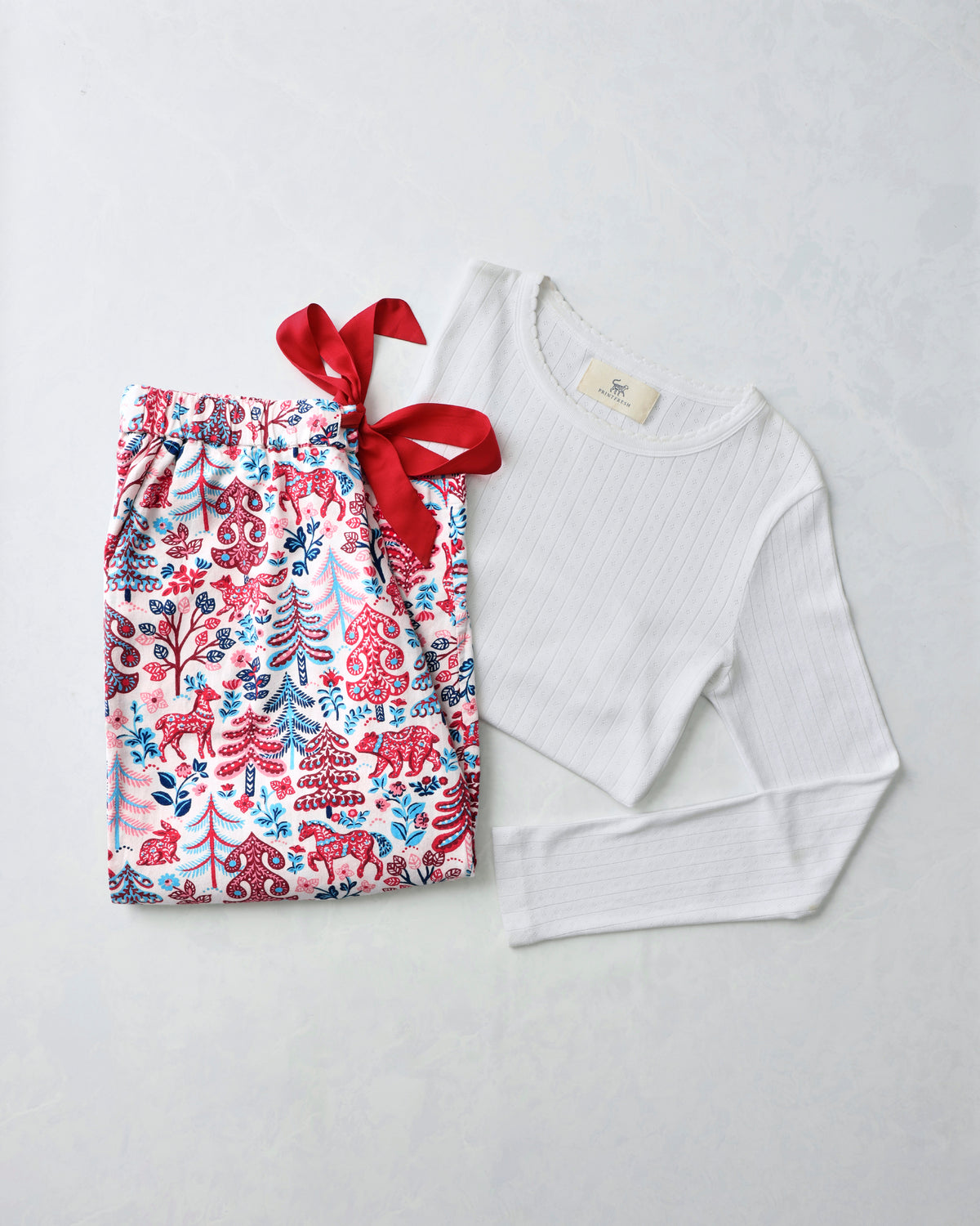 Flat lay of a white pointelle long-sleeve top beside printed pajama pants with red, blue, and green forest motifs and a red ribbon.