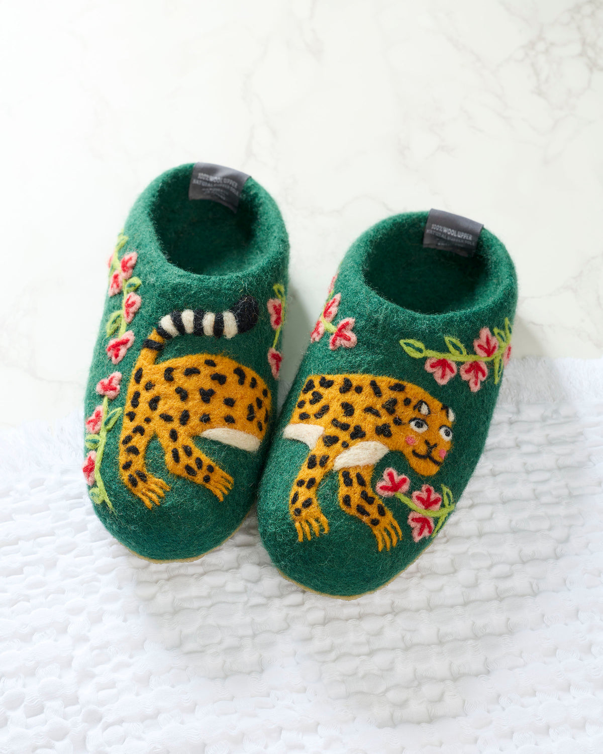 Pair of green slippers with embroidered yellow spotted leopards and pink flowers on a white textured surface.
