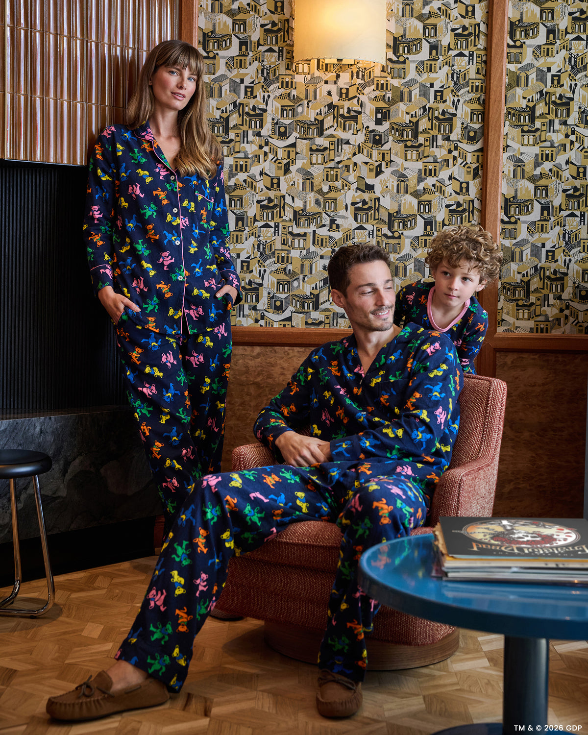 Three people wearing matching navy Dancing Bears long pajama sets in a living room; one person is seated in a chair and two are standing nearby.