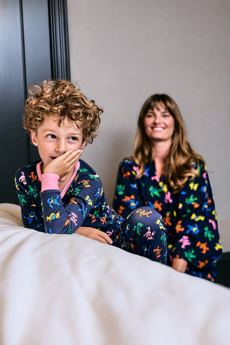 Person with curly hair in patterned pajamas seated on a bed, with another person in matching patterned clothing behind them.
