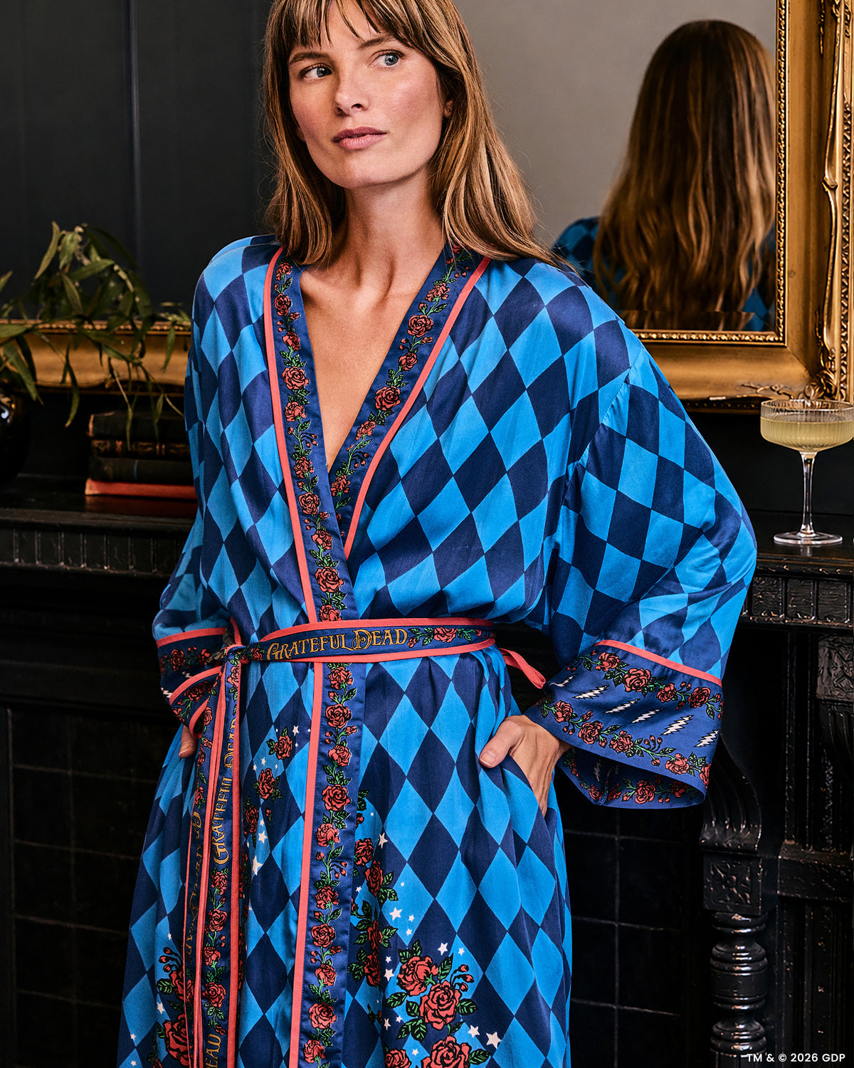 Front three-quarter view of the blue patterned Eco Satin Robe with rose trim and a tied belt, worn in a room with a mirror and cocktail glass nearby.