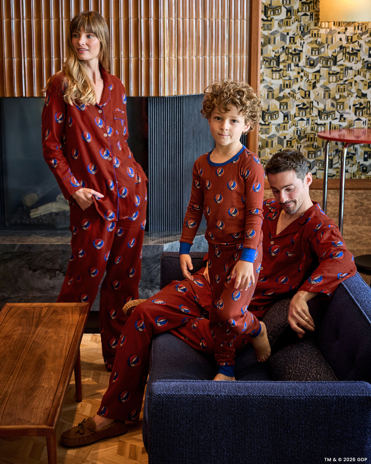 Three people wearing coordinated red patterned pajamas in a living room; one stands while another sits and a child stands in front.