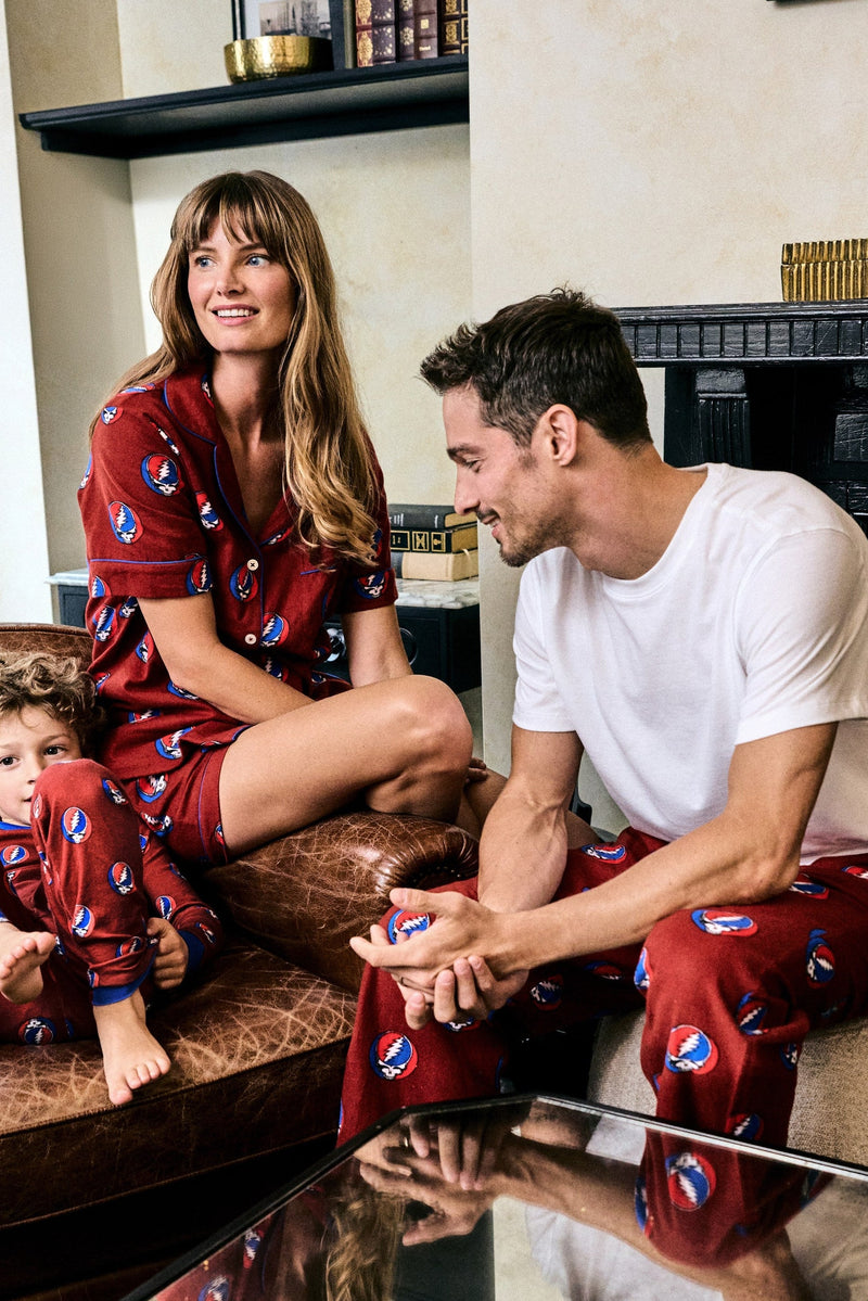 Two people seated indoors, with one wearing a red patterned pajama set and the other wearing a white shirt with red patterned pants.