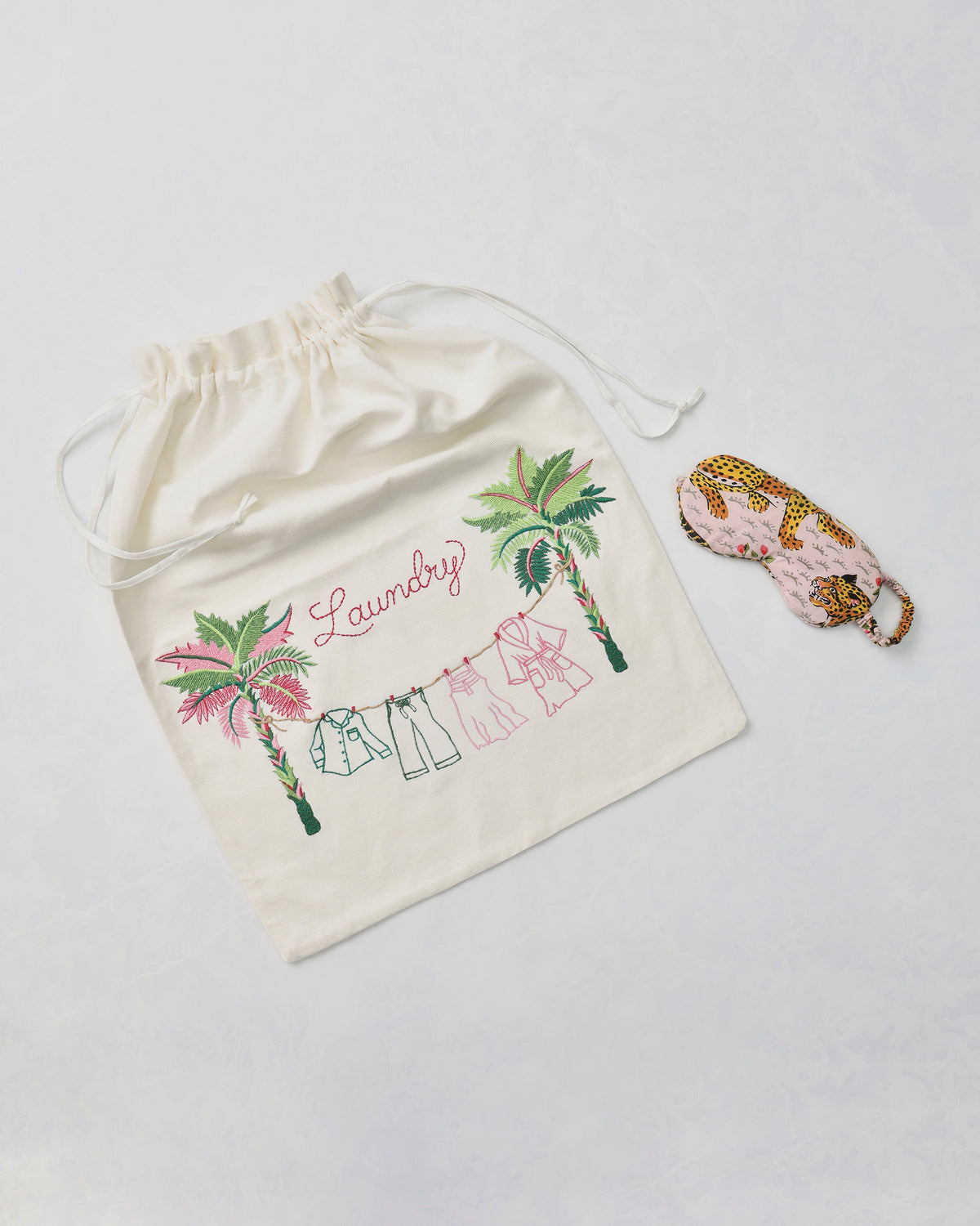 Embroidered drawstring laundry bag with palm trees, clothing line, and the word “Laundry,” beside a leopard-print eye mask.