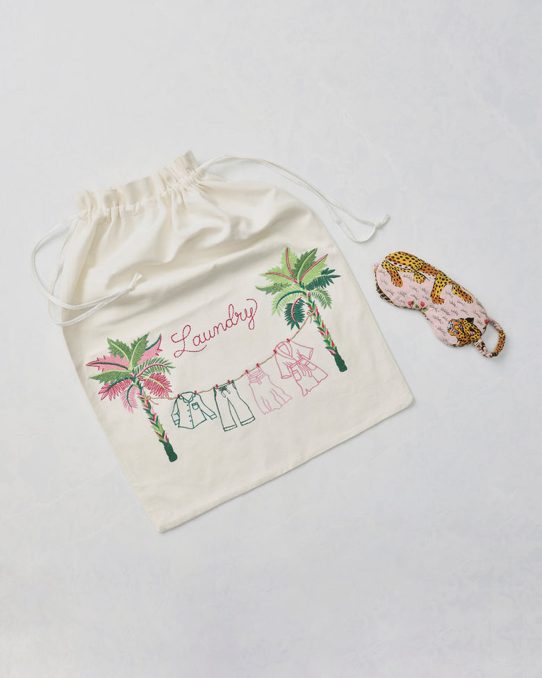Embroidered drawstring laundry bag with palm trees, clothing line, and the word “Laundry,” beside a leopard-print eye mask.