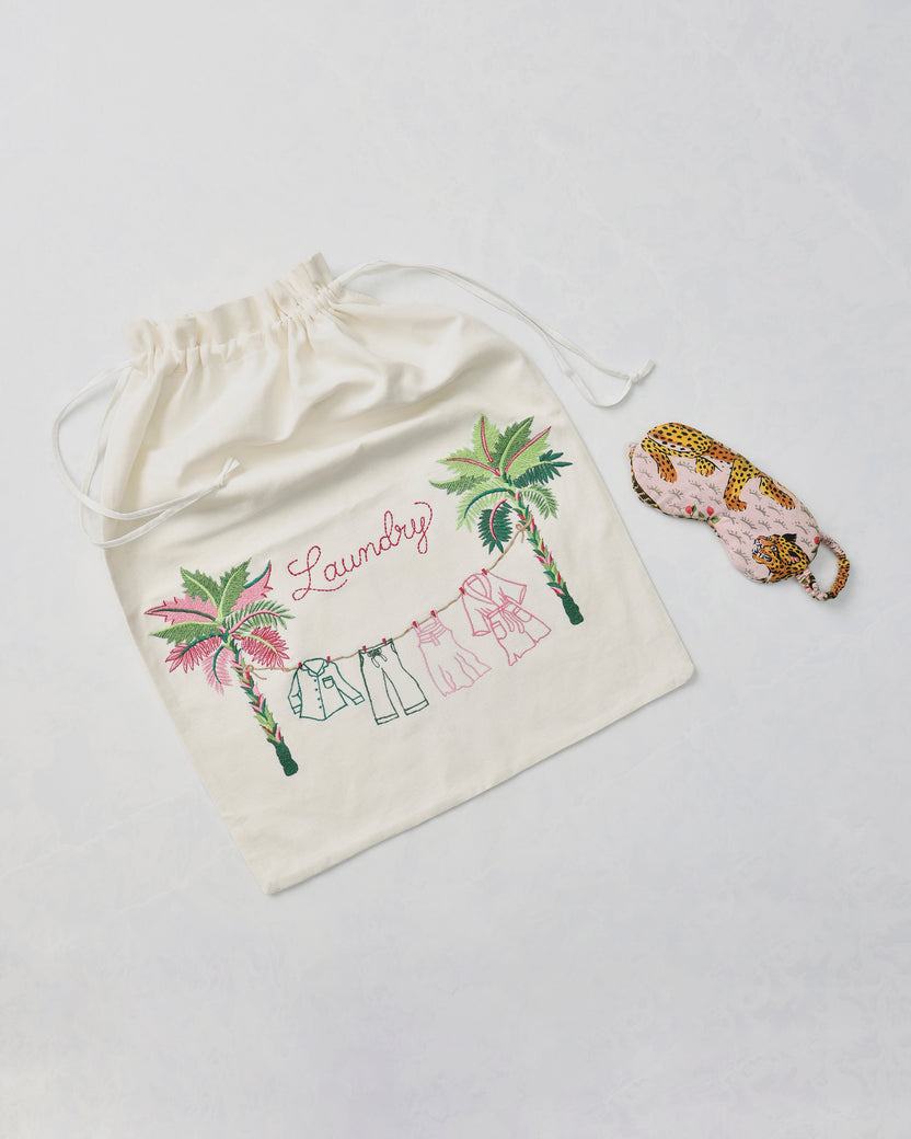Embroidered drawstring laundry bag with palm trees, clothing line, and the word “Laundry,” beside a leopard-print eye mask.