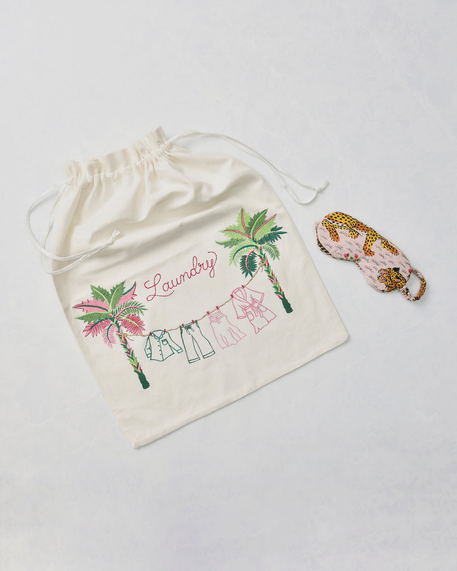 Embroidered drawstring laundry bag with palm trees, clothing line, and the word “Laundry,” beside a leopard-print eye mask.