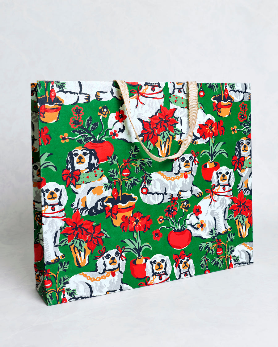 Angled view of the Good Dog Christmas evergreen paper gift bag showing its front and side panels with dogs, poinsettias, ornaments, and tan handles.
