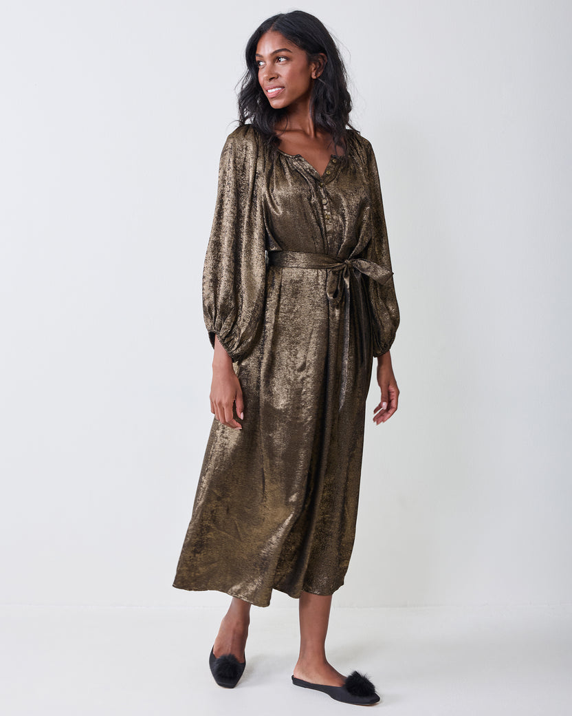 Person wearing a gold long-sleeved dress with a buttoned neckline, waist tie, and flowing skirt.
