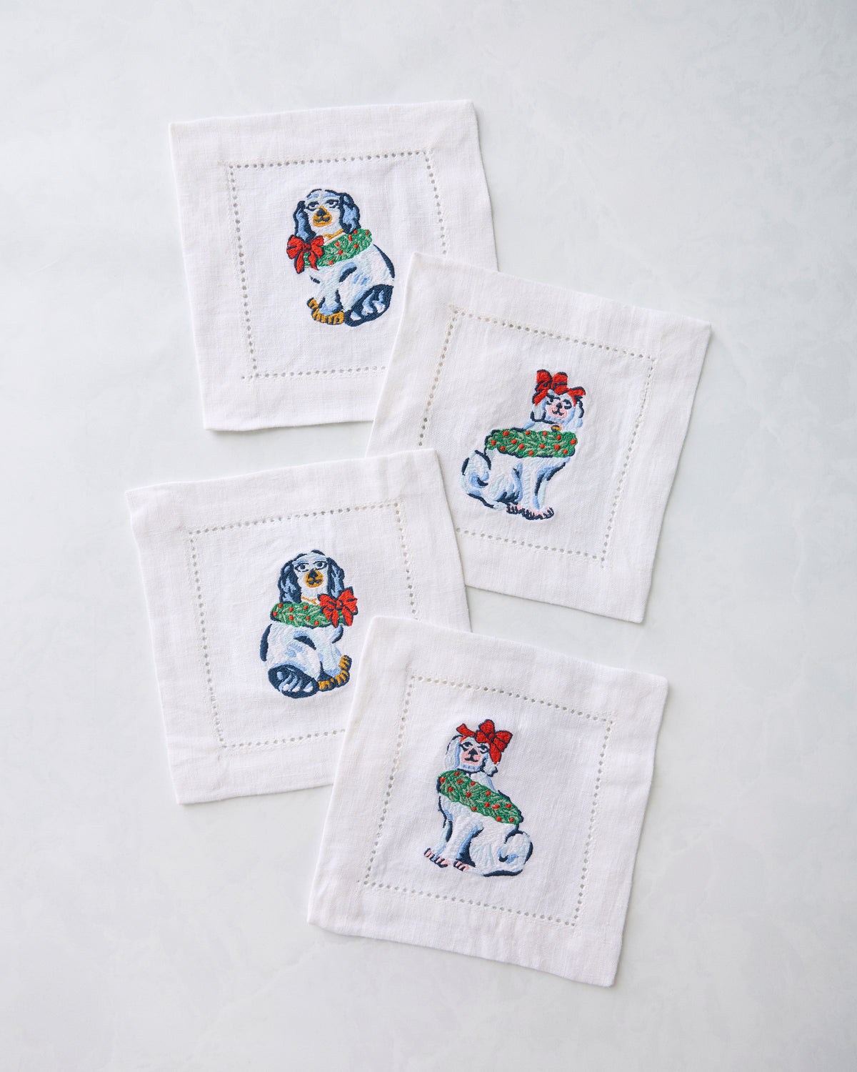 Four white cocktail napkins embroidered with dog figures wearing holiday decorations.
