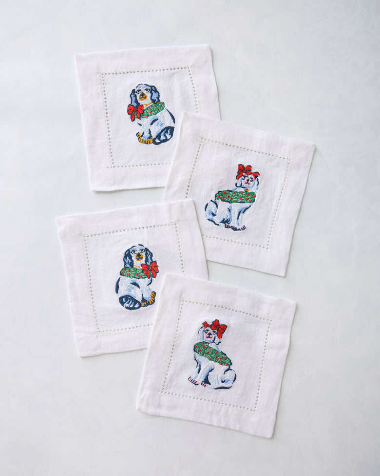 Four white cocktail napkins embroidered with dog figures wearing holiday decorations.