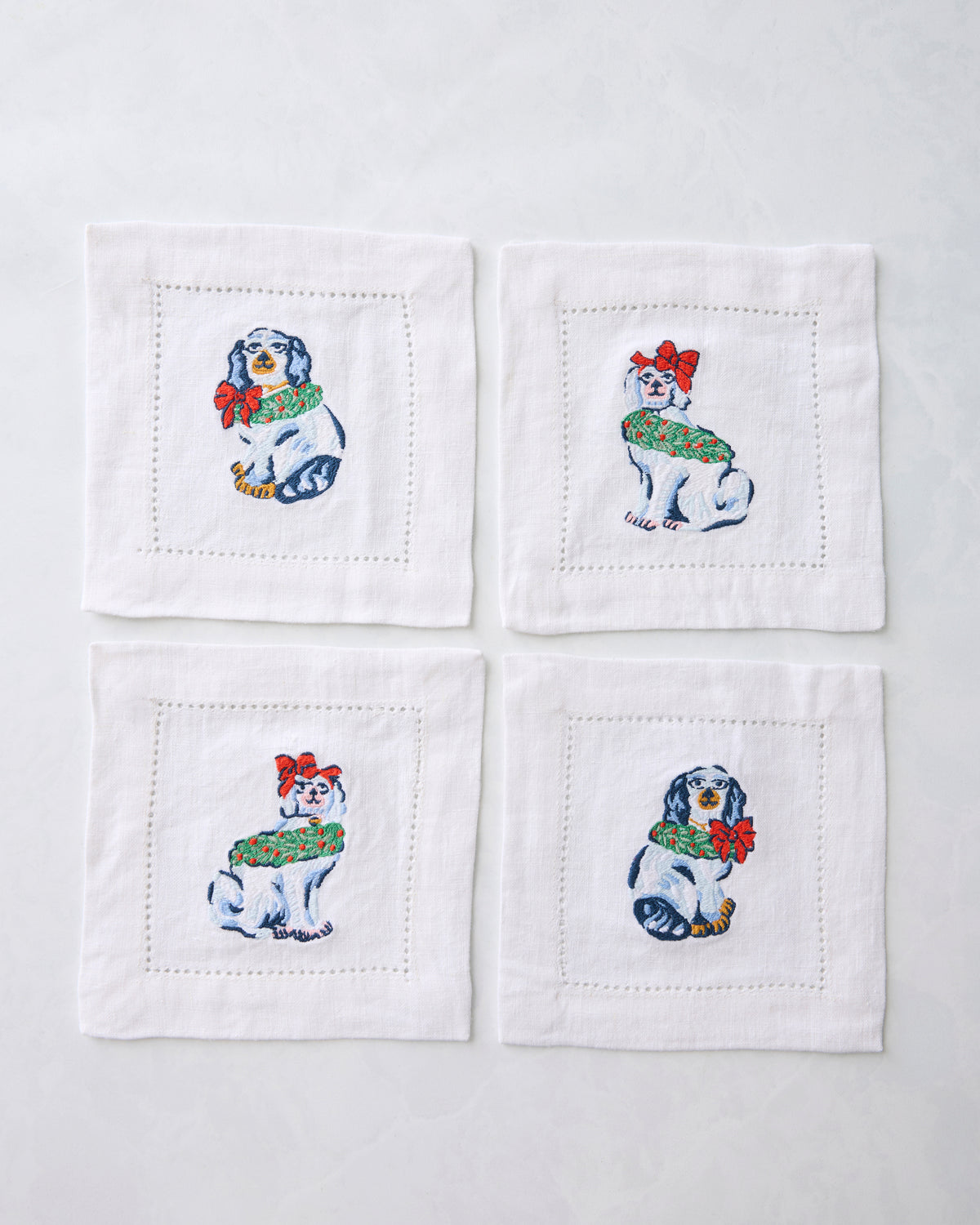 Four white cocktail napkins arranged in a square, each embroidered with a Christmas dog wearing a green wreath and red bow.