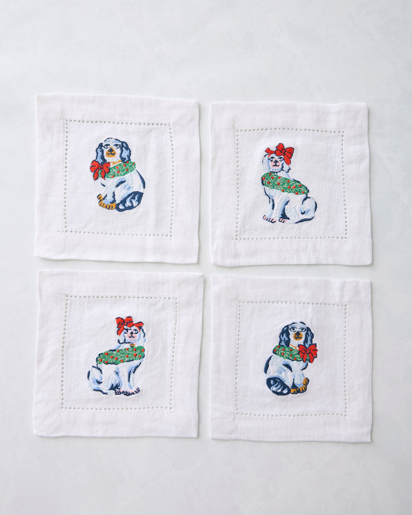 Four white cocktail napkins arranged in a square, each embroidered with a Christmas dog wearing a green wreath and red bow.