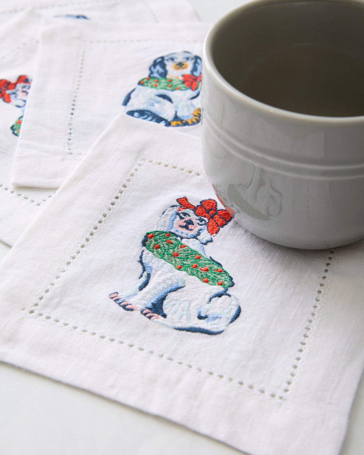 White cocktail napkin embroidered with a Christmas dog, shown beneath a white cup with other napkins in the background.