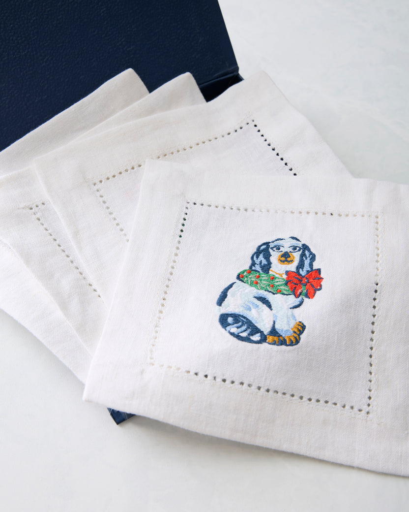 Close view of a white cocktail napkin embroidered with a Christmas dog wearing a green wreath and red bow.