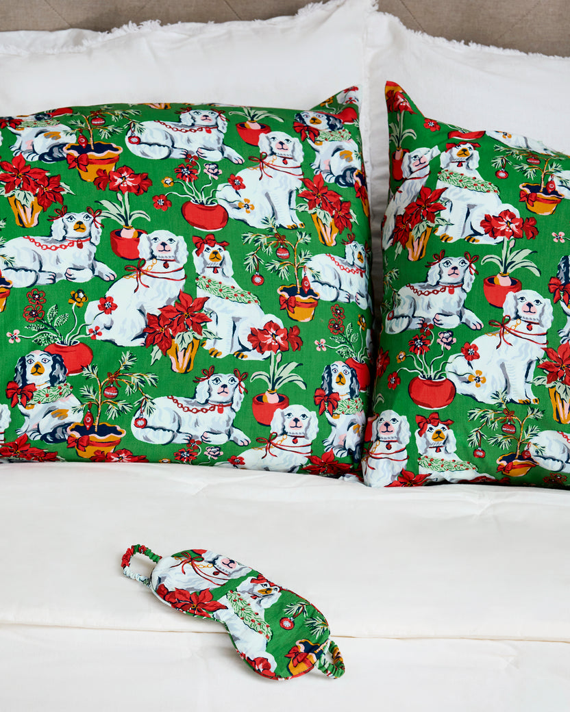 Two evergreen pillowcases with a repeating print of white dogs, red poinsettias, potted plants, and holiday decorations, arranged on a bed.