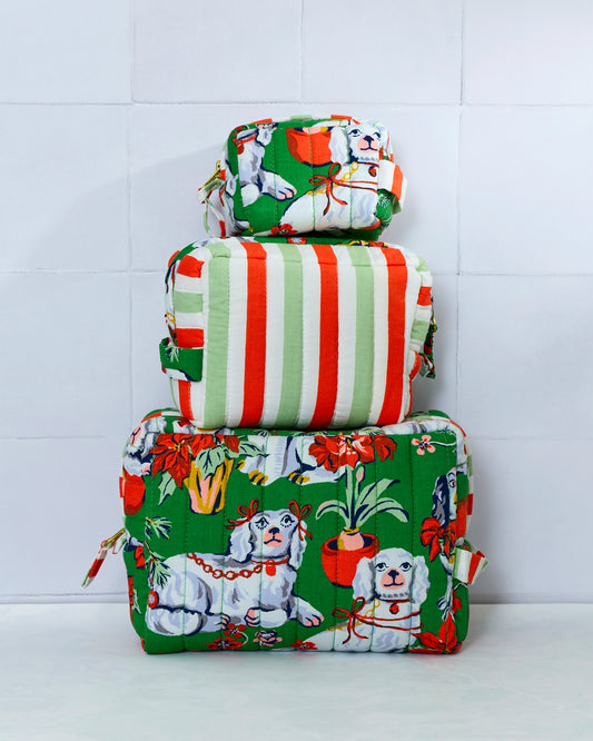 Three quilted evergreen Good Dog Christmas pouches stacked by size, showing white dogs, potted plants, holiday flowers, and the striped back of the middle pouch.