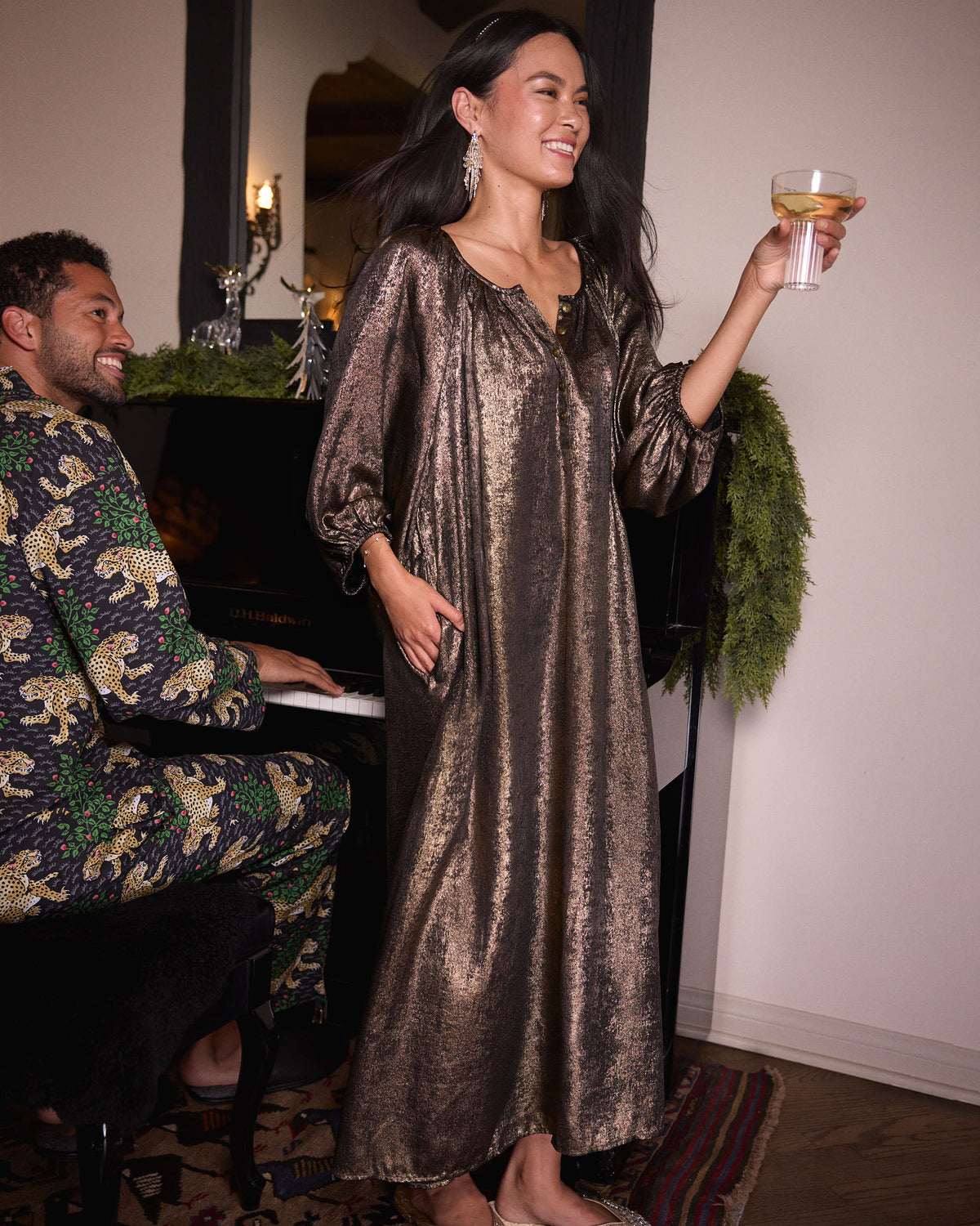Person wearing the gold Glitzy Lamé Felicity dress and holding a drink beside a piano.
