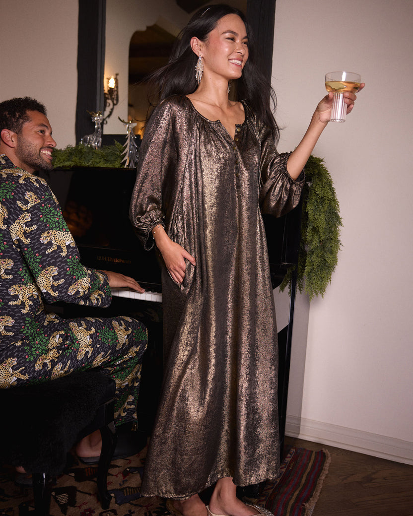 Person wearing the gold Glitzy Lamé Felicity dress and holding a drink beside a piano.