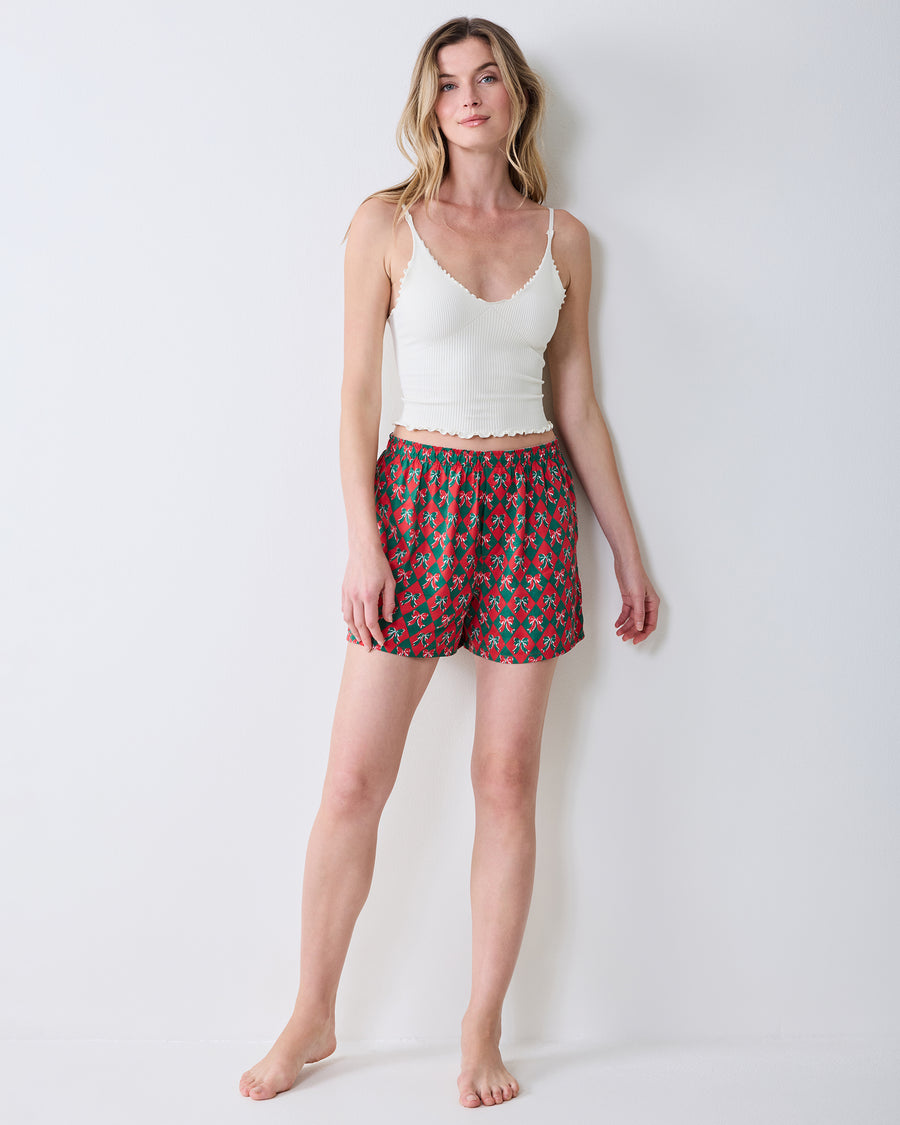 Full-length front view of red boxer shorts with a green argyle and bow print, styled with a white camisole.
