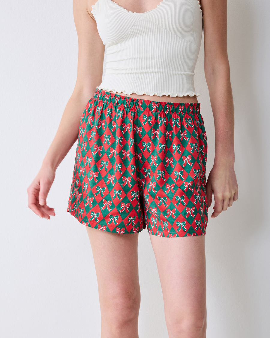 Close front view of red boxer shorts with a green argyle and bow print, showing the elastic waistband and leg openings.