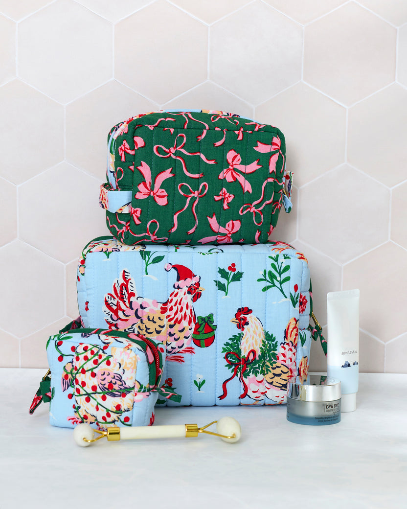 Three quilted pale blue and green pouches from the Holiday Hens set stacked with skincare items and a facial roller; the largest pouch features illustrated hens and holiday greenery.