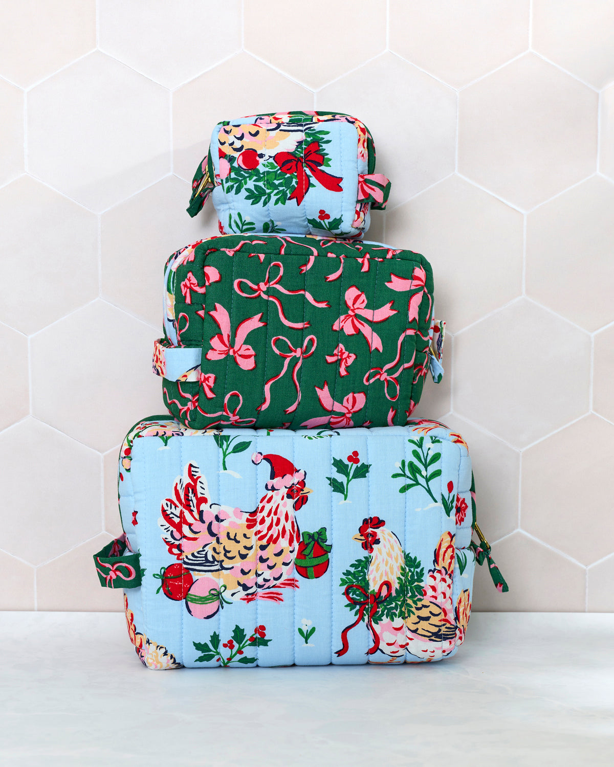 Three quilted pouches from the pale blue Holiday Hens set stacked by size, showing hen, greenery, bow, and holiday illustrations.