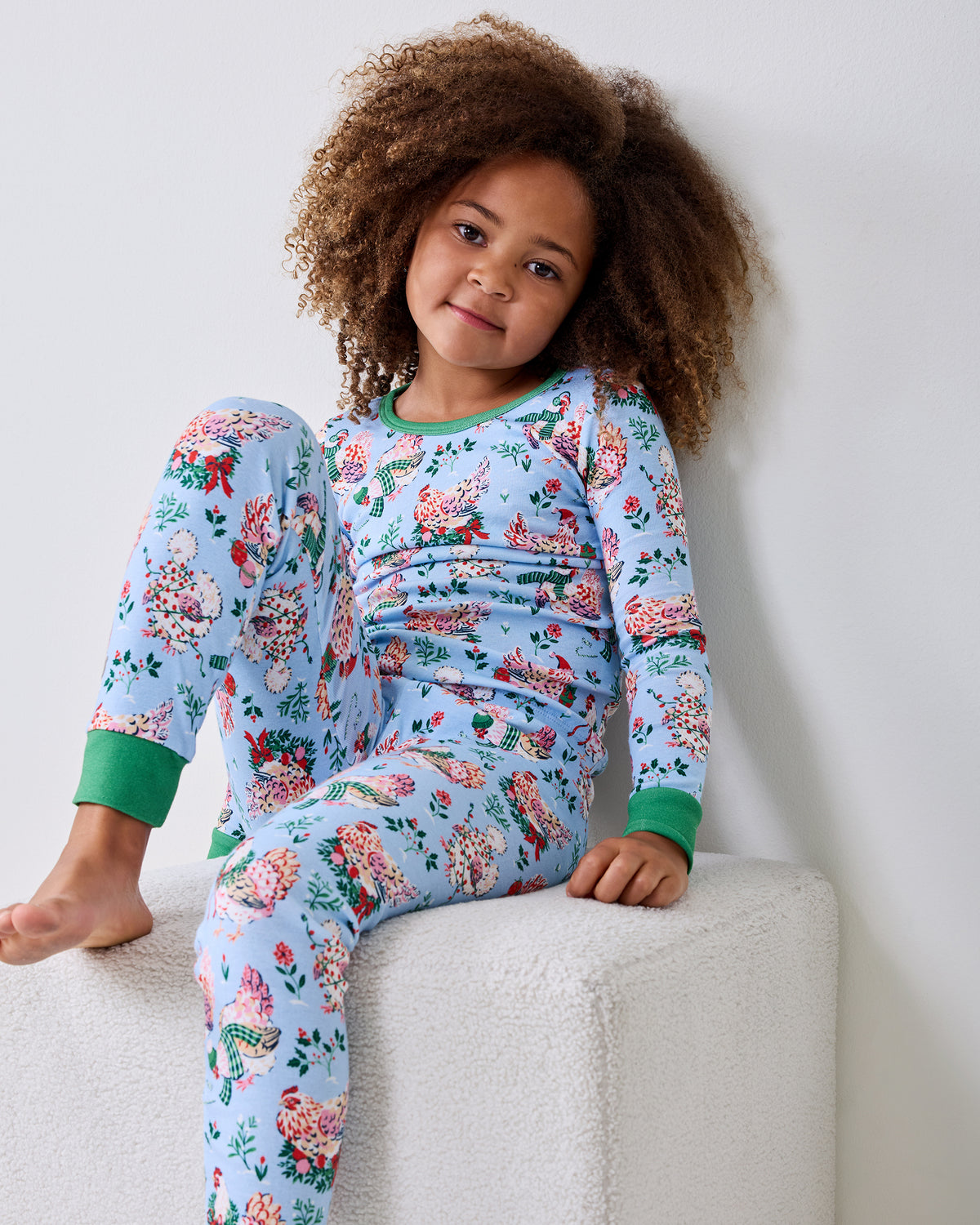 Child wearing a pale blue long-sleeve Holiday Hens pajama set with a floral and hen print, seated on a white ottoman.