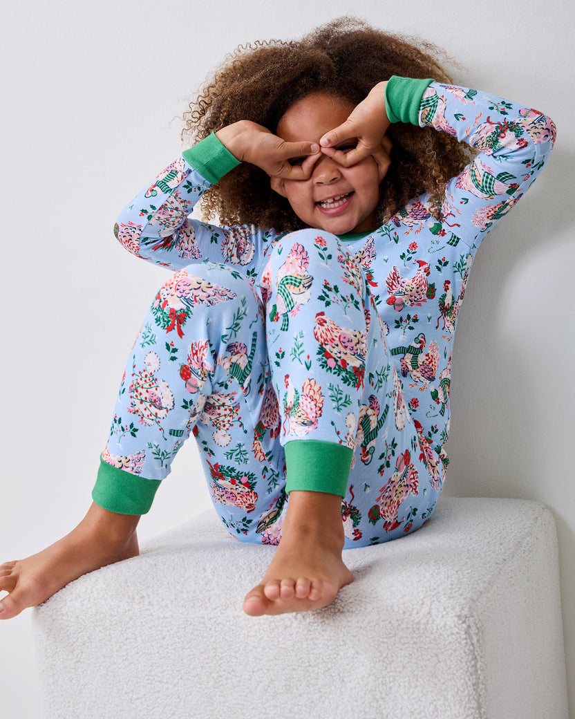 Child wearing a pale blue Holiday Hens pajama set sits on a white ottoman with both hands over their eyes.