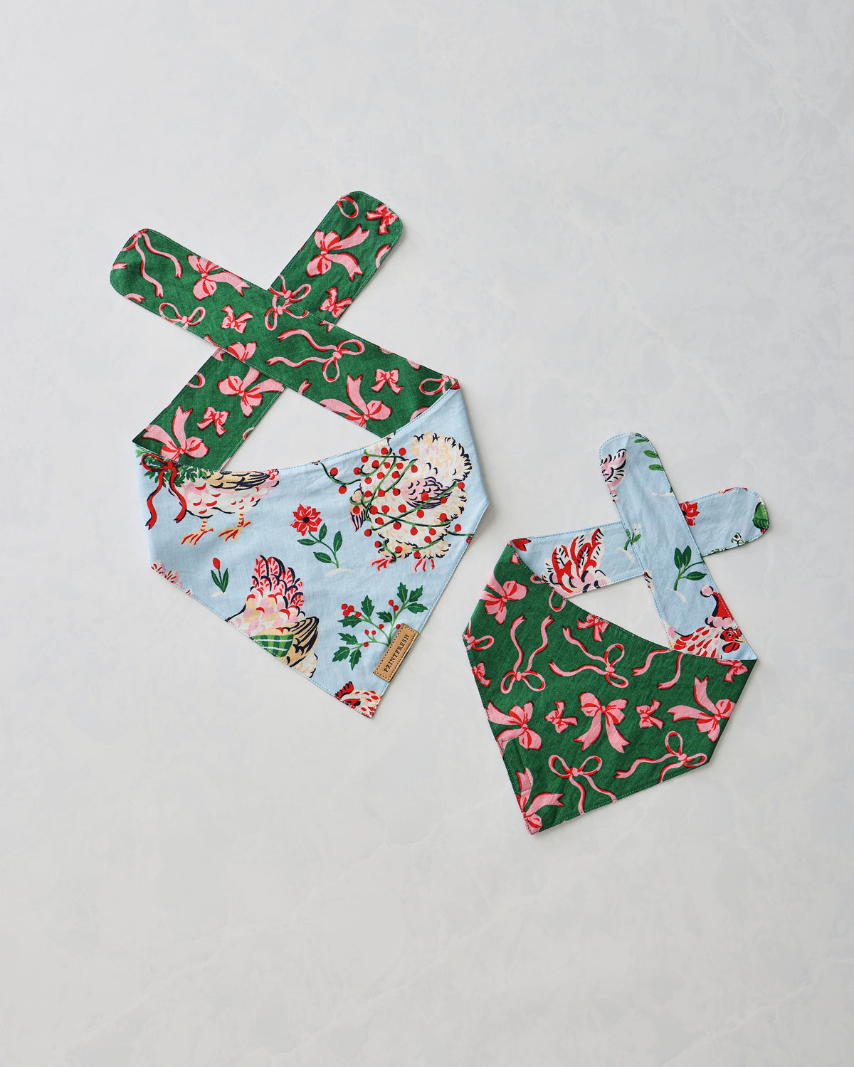 Two pet bandanas displayed flat: one pale blue with floral and hen print and one green with pink bow print.