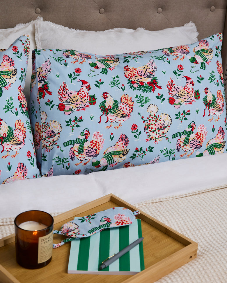 Two pale blue Holiday Hens pillowcases displayed on a bed beside a tray with a candle, notebook, and pen.
