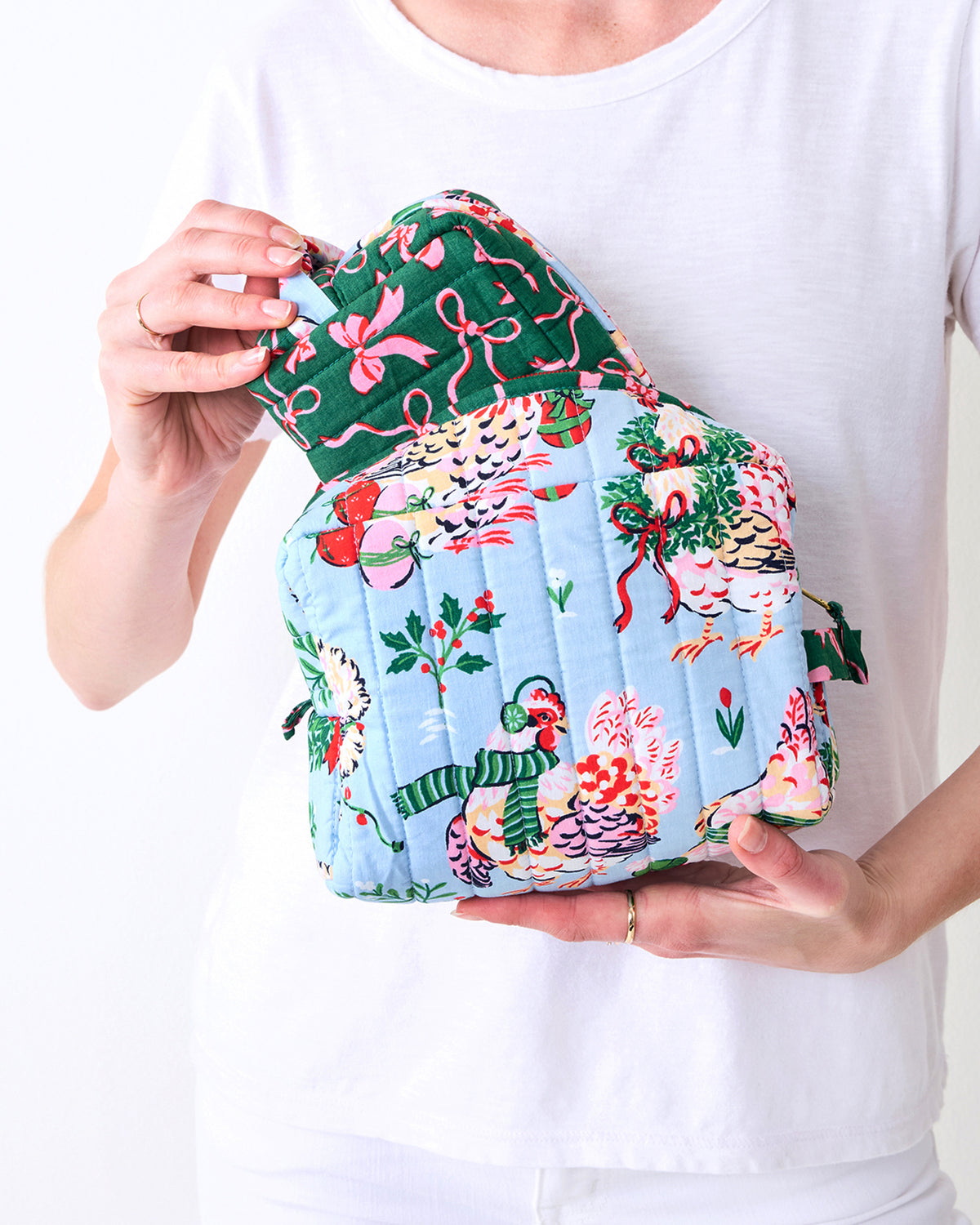 Person holding the largest pale blue Holiday Hens pouch with the green and pink bow-print pouch resting on top, showing the hen and holiday illustrations.