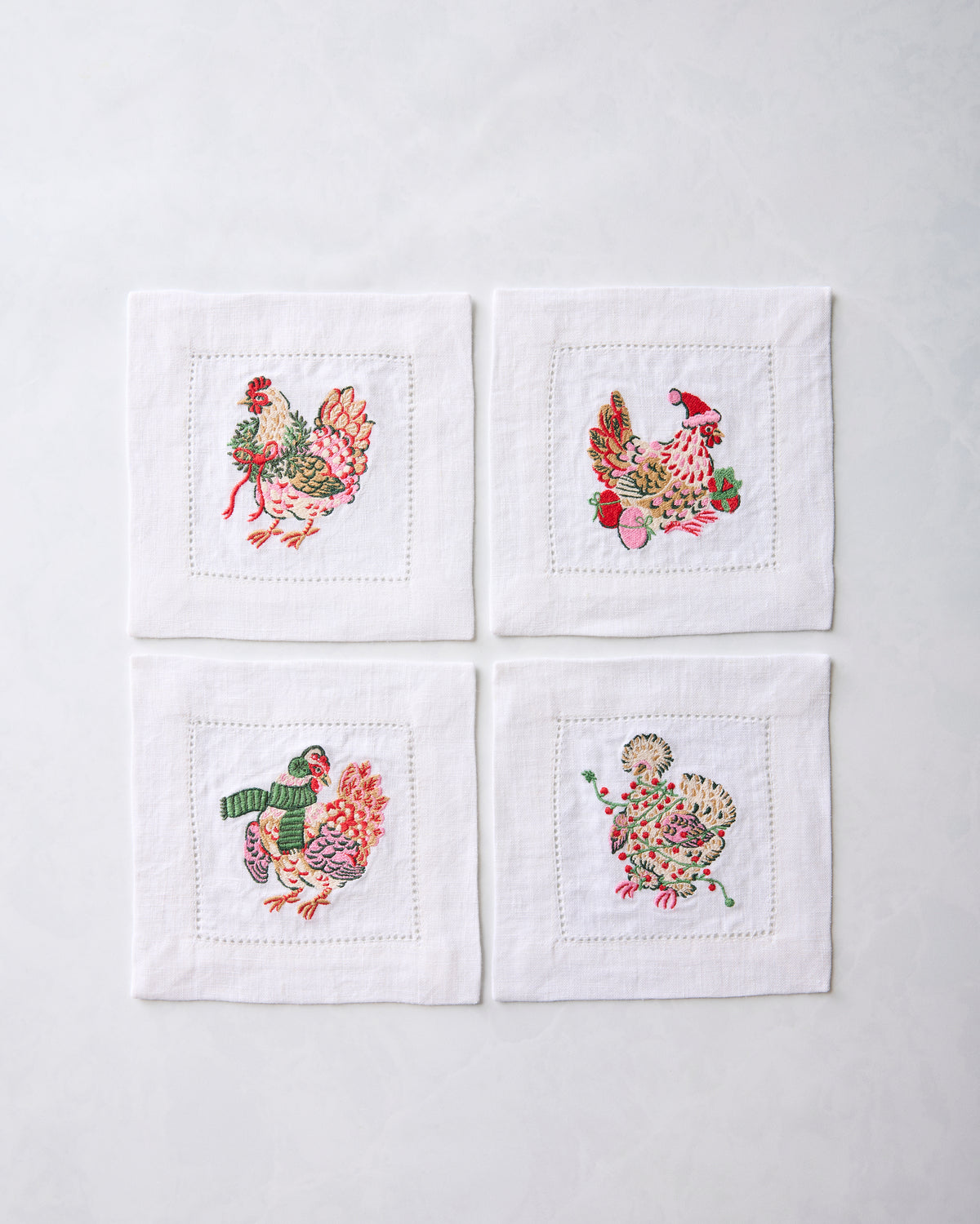 Set of four white cocktail napkins arranged in a square, each embroidered with a different holiday chicken