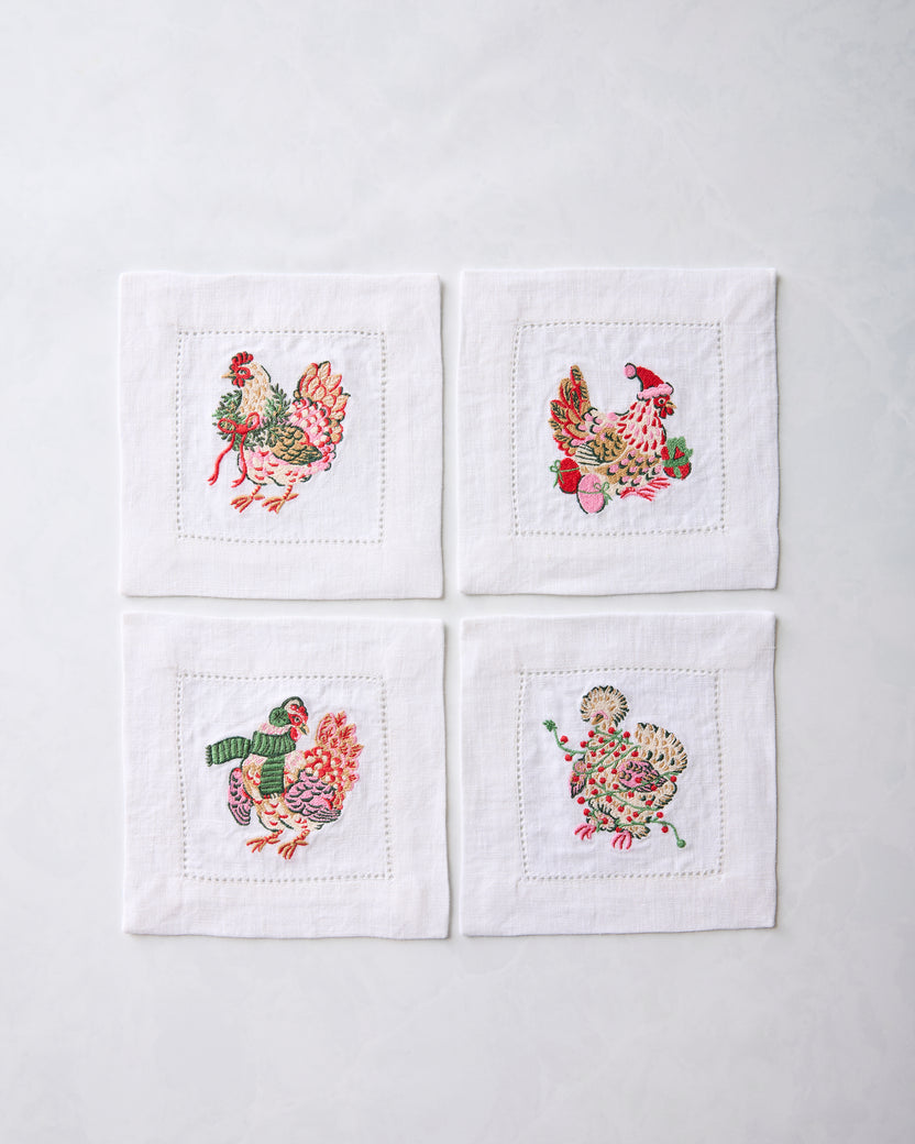 Set of four white cocktail napkins arranged in a square, each embroidered with a different holiday chicken