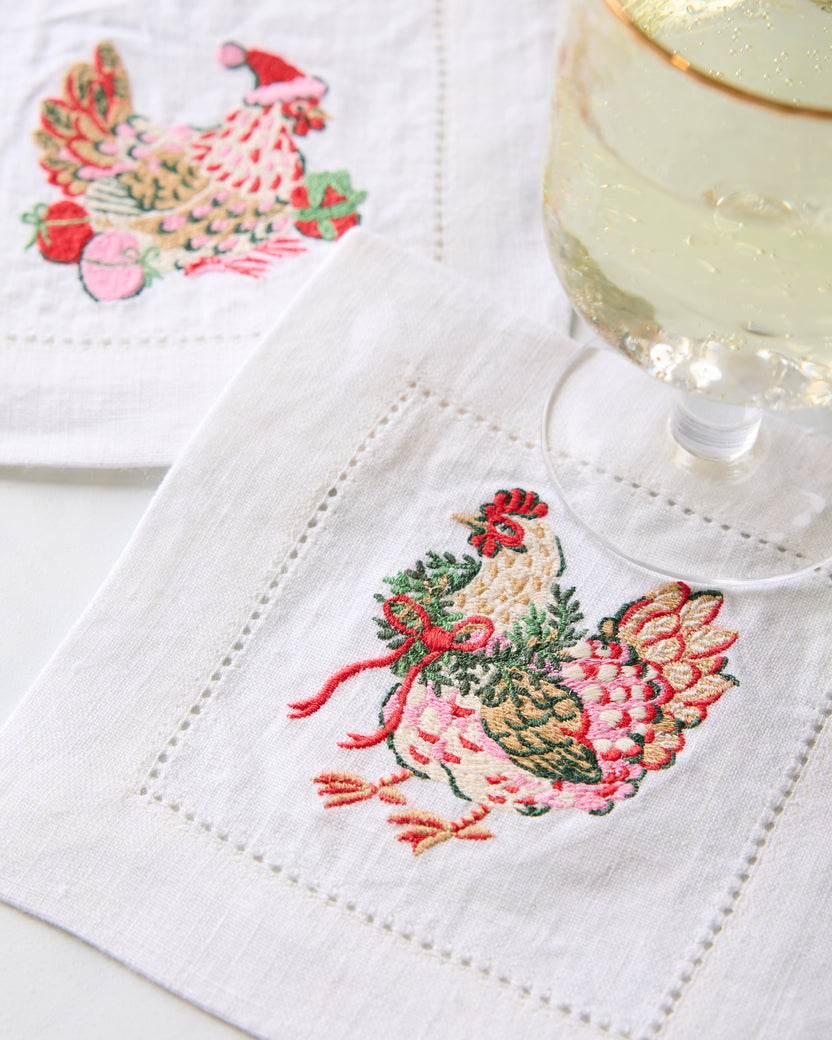 Close-up of an embroidered chicken cocktail napkin beside a stemmed glass