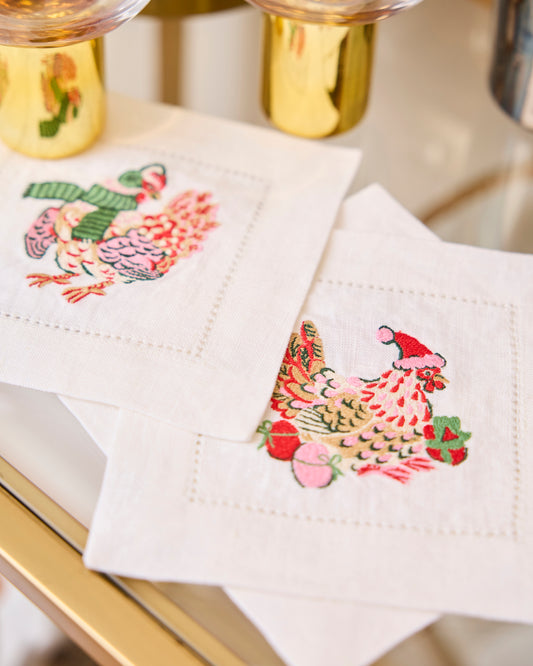 Embroidered chicken cocktail napkins arranged on a table beside stemmed glasses