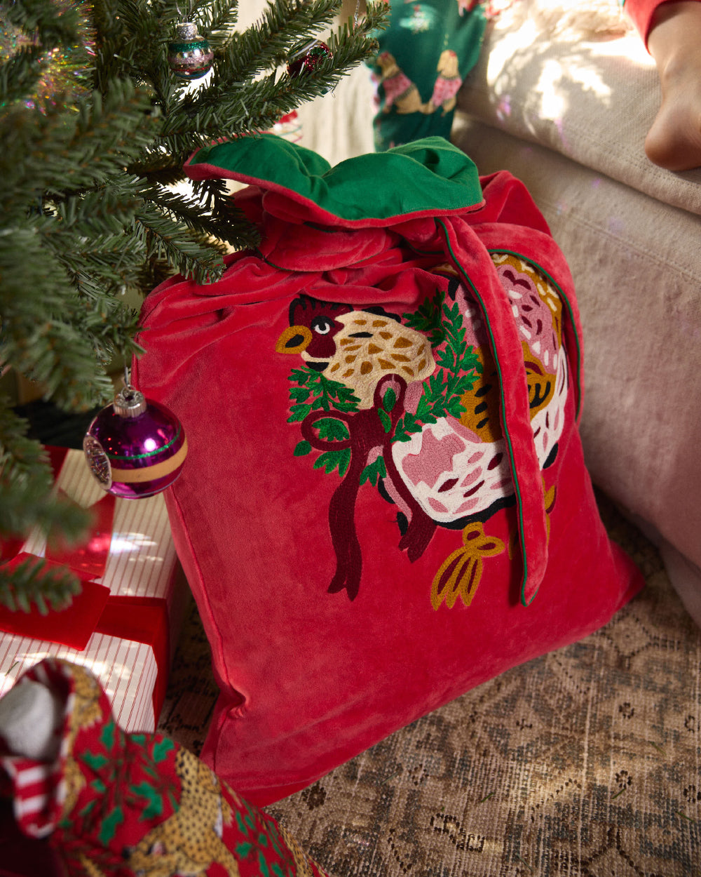 Red embroidered gift sack with a tie, decorated with holiday hens and greenery beside a Christmas tree.