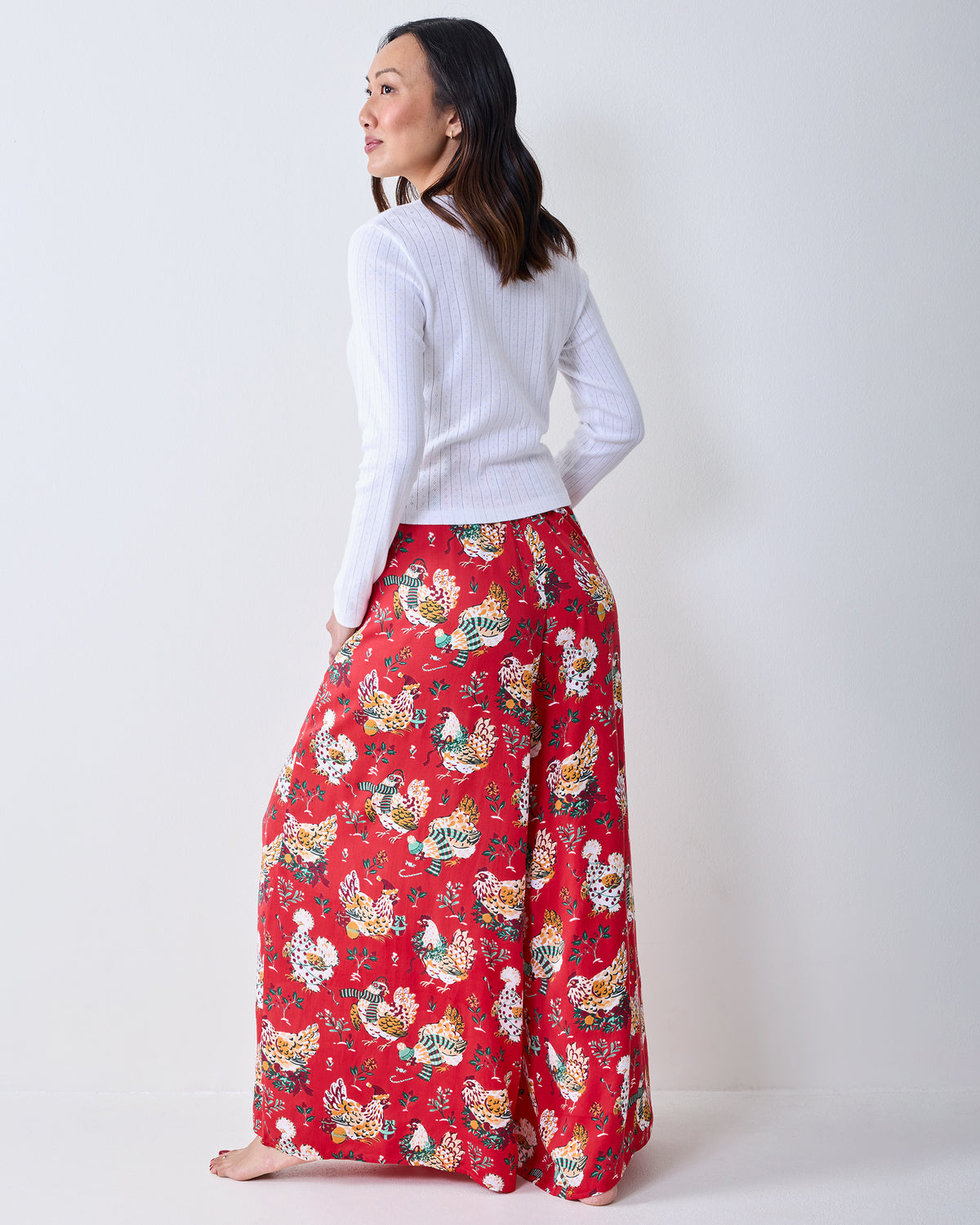 Back view of a model wearing red Holiday Hens wide-leg pants with an all-over chicken and floral print.