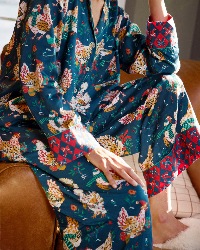 Close view of a navy pajama top and pants with festive hen print, white piping, and red patterned cuffs.